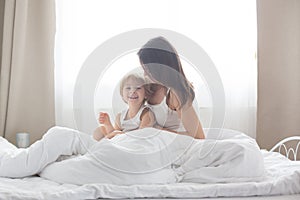 Beautiful blond child and his mother, playing at home in the morning in bed, smiling, laughing and having fun