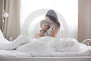 Beautiful blond child and his mother, playing at home in the morning in bed, smiling, laughing and having fun