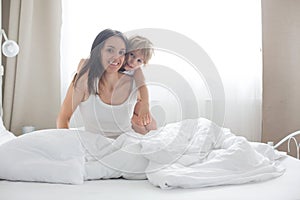 Beautiful blond child and his mother, playing at home in the morning in bed, smiling, laughing and having fun