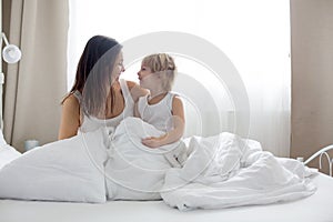 Beautiful blond child and his mother, playing at home in the morning in bed, smiling, laughing and having fun