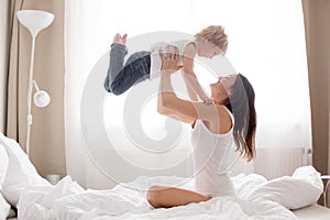 Beautiful blond child and his mother, playing at home in the morning in bed, smiling, laughing and having fun