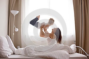 Beautiful blond child and his mother, playing at home in the morning in bed, smiling, laughing and having fun