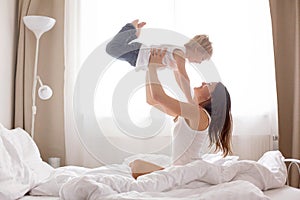 Beautiful blond child and his mother, playing at home in the morning in bed, smiling, laughing and having fun