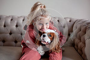 Beautiful blond girl child sits at home on a sofa, playing with and hugging dog, creating heartwarming moments of joy