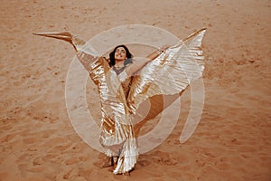 A beautiful girl in a golden with brilliant dress with wings, suit is dancing an oriental, East dance in the desert