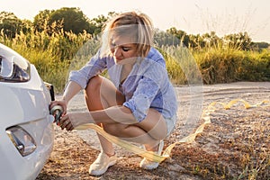 Beautiful blond girl is holding a tow rope on a broken car on a rural road in the rays of the sunset