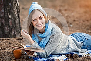 Beautiful blond girl resting in spring or autumn forest read book and, lie with cup of tea and book. Confident caucasian