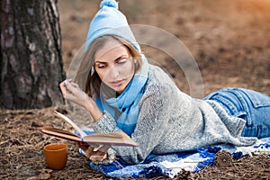 Beautiful blond girl resting in spring or autumn forest read book and, lie with cup of tea and book. Confident caucasian