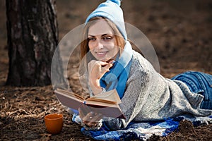 Beautiful blond girl resting in spring or autumn forest read book and, lie with cup of tea and book. Confident caucasian