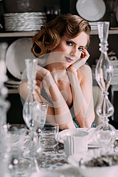 Beautiful blond girl with vintage make up and hairstyle in evening shining dress sitting at table with plates and glasses