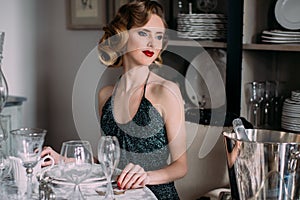 Beautiful blond girl with vintage make up and hairstyle in evening shining dress sitting at table with plates and glasses