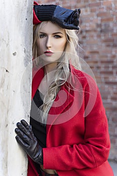 A beautiful blond girl wearing a red coat and gloves stands by a ruined wall and brick arch near an old building. Fashionable,