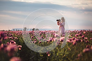 Beautiful blond-hair woman in hat takes photos in the Lilac Poppy Flowers field