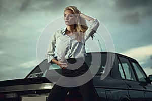 Beautiful blond lady in black striped high waisted pants and oversized white blouse standing at her old car looking away