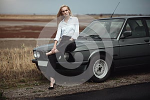 Beautiful blond lady in black striped high waisted pants, white blouse and high heeled shoes sitting on the hood of her old car photo