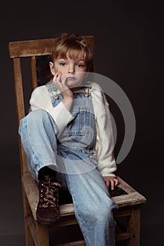 Beautiful blond little girl sitting on the old chair with sad expression on her face. Her foot in trendy boot up on the chair