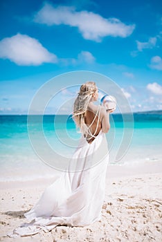 Beautiful blond long hair fiancee in a long white open back wedding dress stand on the white sand beach with a pearl. Looking to