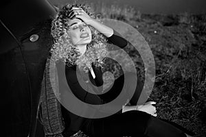 Beautiful blond smiling woman with afro curly hair wearing black trouser suit sitting on the ground leaning on her car