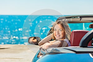 Beautiful blond smiling young woman in convertible top automobile looking sideways while parked near ocean waterfront photo