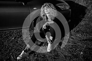 Beautiful blond woman with afro curly hair wearing black trouser suit and white sneakers sitting on the ground leaning on her car