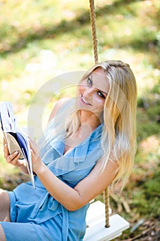 Beautiful blond woman in blue dress reading a book on a swing