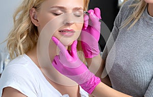 Beautiful blond woman with curly hair having Permanent Make-up Tattoo on her Eyebrows. Eyelash artist plucks eyebrows with