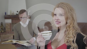 Beautiful blond woman drinking coffee in the foreground while modestly dressed man reading the book on the background