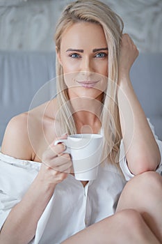 Beautiful blond woman drinking a coffee while sitting on her bed at home