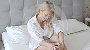 Beautiful blond woman drinking a coffee while sitting on her bed at home