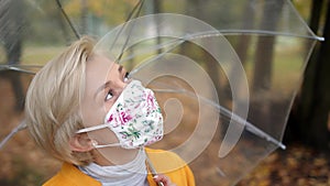 Beautiful blond woman with face mask holding transparent umbrela in the park in autumn. Coronavirus outbreak 2020