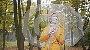 Beautiful blond woman with face mask spinning transparent umbrela in the park in autumn. Coronavirus outbreak 2020