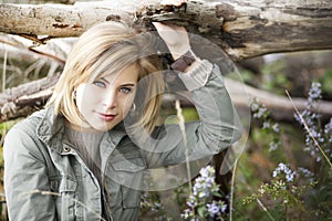 A beautiful blond woman holding onto a tree limb while sitting amonst flowers.