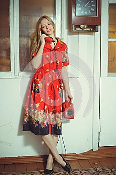 Beautiful blond woman with an old wired phone dressed in a red dress, standing in an old house, vintage style