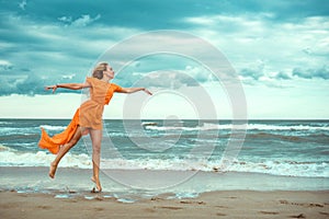 Beautiful blond woman in orange mini dress with flying train dancing barefoot on the wet sand at the storming sea