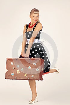 Beautiful blond woman in pinup style dressed in a polka-dot dress is standing and flirtatiously holding a brown suitcase, white