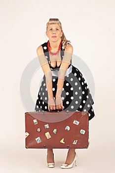 Beautiful blond woman in pinup style, dressed in a polka-dot dress, stands and hardly holds a heavy suitcase, white background