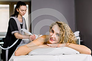 Beautiful blond woman smiling, looking at camera while getting ultrasound cavitation treatment by cosmetologist. Anti