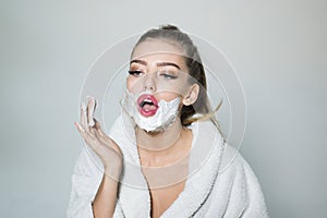 Beautiful blond woman in white bathrobe on gray background. Pretty girl applying shaving foam on her cheeks