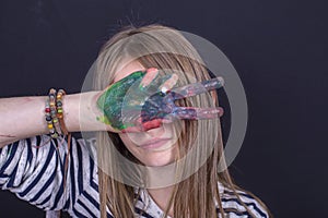 Beautiful blond young girl with freckles and hands painted in colorful paints indoors on black background, closeup portrait