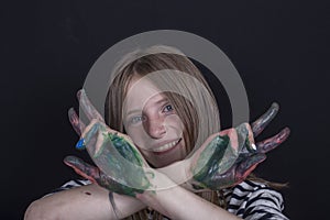 Beautiful blond young girl with freckles and hands painted in colorful paints indoors on black background, closeup portrait