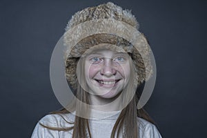 Beautiful blond young girl with a winter fur hat indoors on a black background, closeup portrait