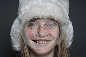 Beautiful blond young girl with a winter fur hat indoors on a black background, closeup portrait