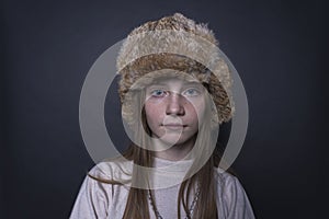 Beautiful blond young girl with a winter fur hat indoors on a black background, closeup portrait