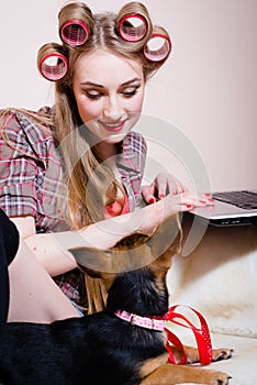 Beautiful blond young pinup woman having fun playing with cute small dog relaxing lying in bed typing on laptop