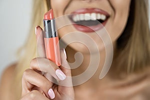 Beautiful blond young smiling woman holding lipstick in hand close up isolated white background