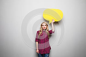 Beautiful blond young woman holding yellow blank speech bubble over grey background