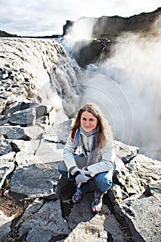 Beautiful blond young woman sitting on stones near Dettifoss waterfall, Iceland