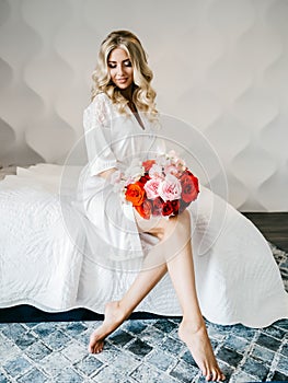 Beautiful blonde with a bouquet of flowers sits on a bed in a white room