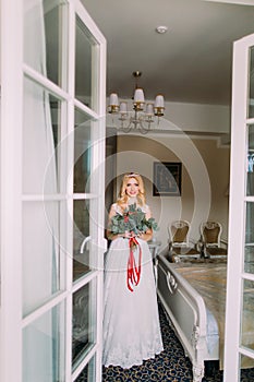 Beautiful blonde bride with bouquet of red roses standing near the window