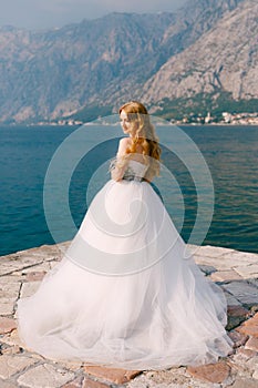 A beautiful blonde bride in a puffy white dress stands on a pier in the Bay of Kotor
